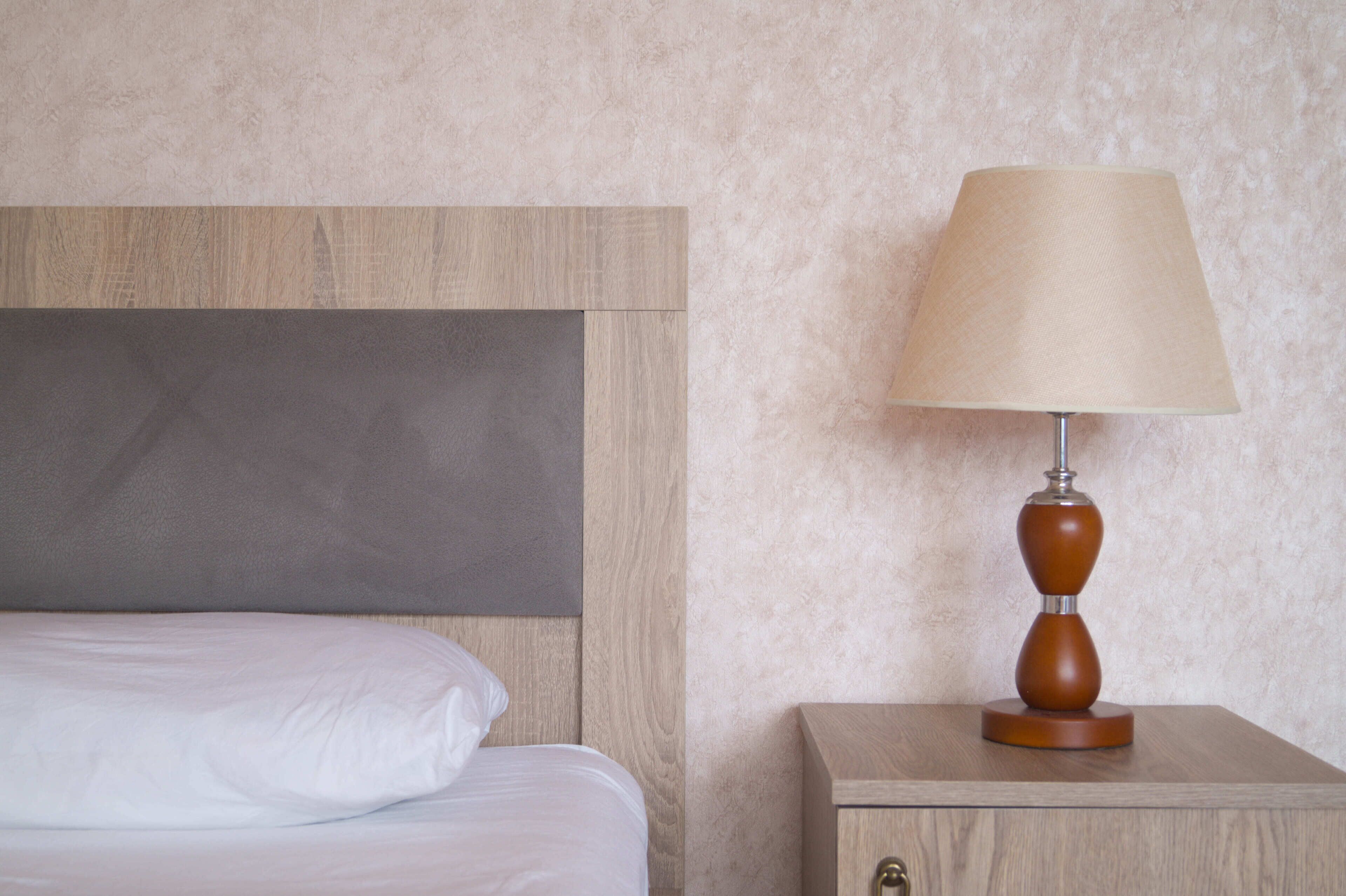 Ein ordentlich bezogenes Bett mit weißen Leinen, ein Nachttisch aus Holz mit einer Lampe und eine strukturierte Wand.