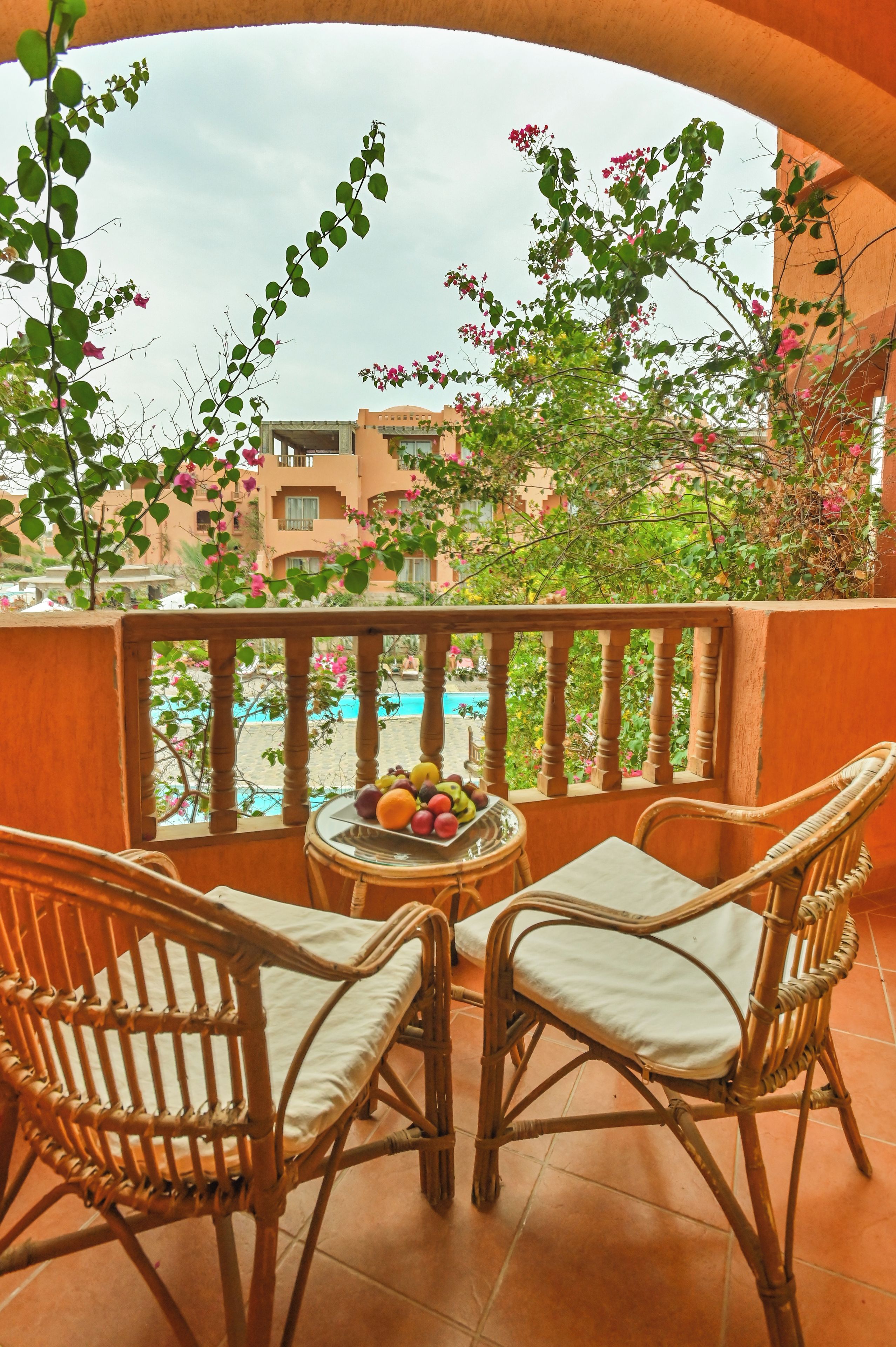 A balcony with wicker chairs, a fruit bowl, and a view of a swimming pool and buildings.