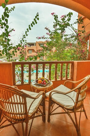 Een balkon met rieten stoelen, een schaal fruit en uitzicht op een zwembad en gebouwen.