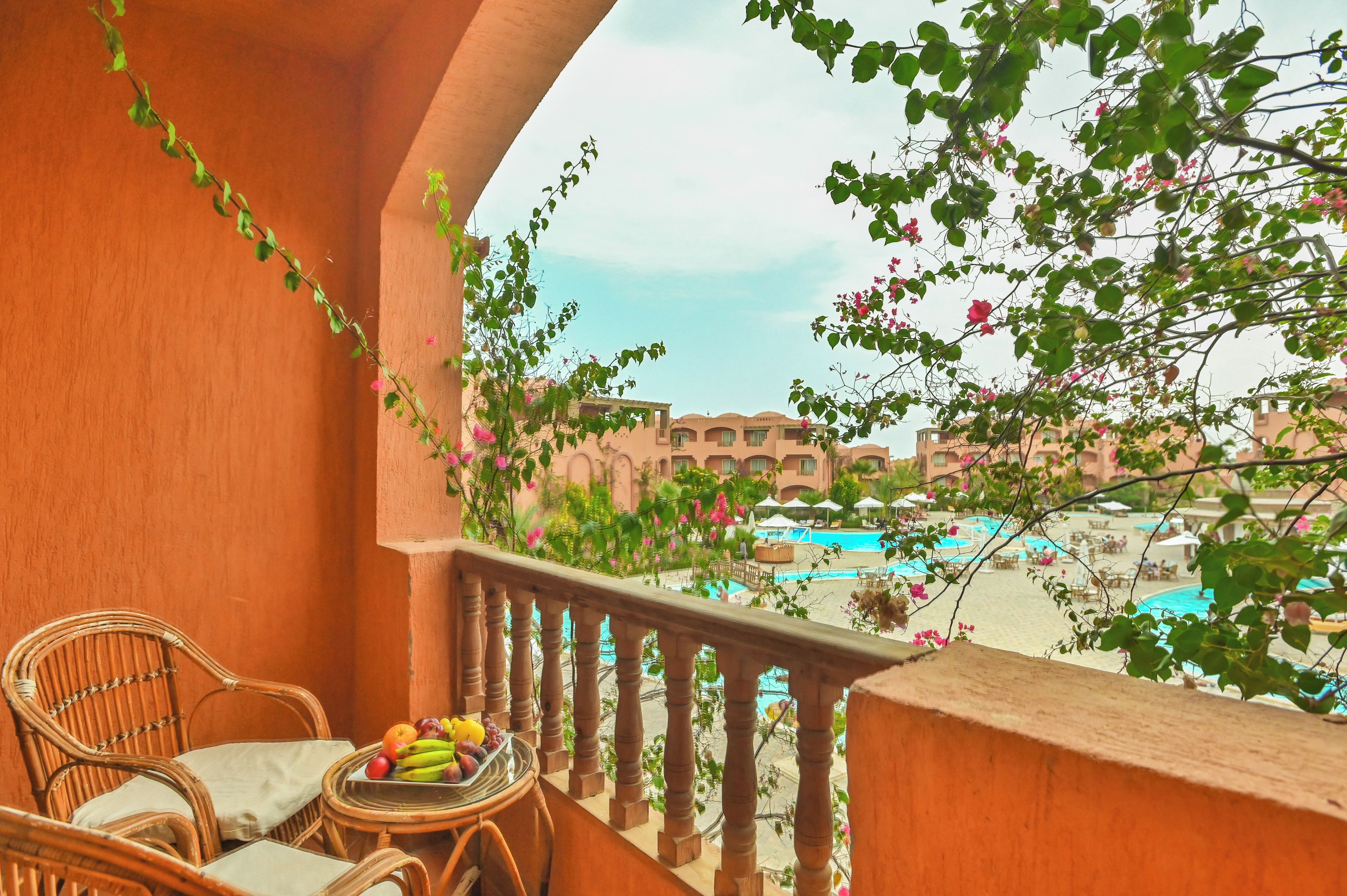A balcony with wicker chairs and a table with fruit, overlooking a pool and buildings.