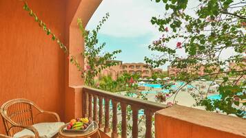A balcony with wicker chairs and a table with fruit, overlooking a pool and buildings.