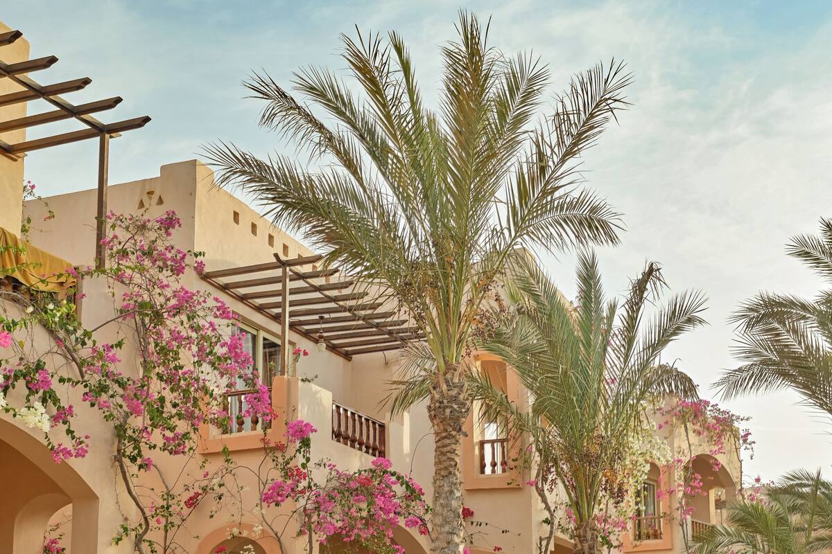 Un bâtiment traditionnel doté d’arches au niveau des fenêtres et de balcons, entouré d’une végétation luxuriante et de palmiers.