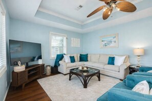Un salon avec un canapé d’angle beige, une table basse en verre, un fauteuil bleu, un meuble de rangement en bois et un ventilateur de plafond.
