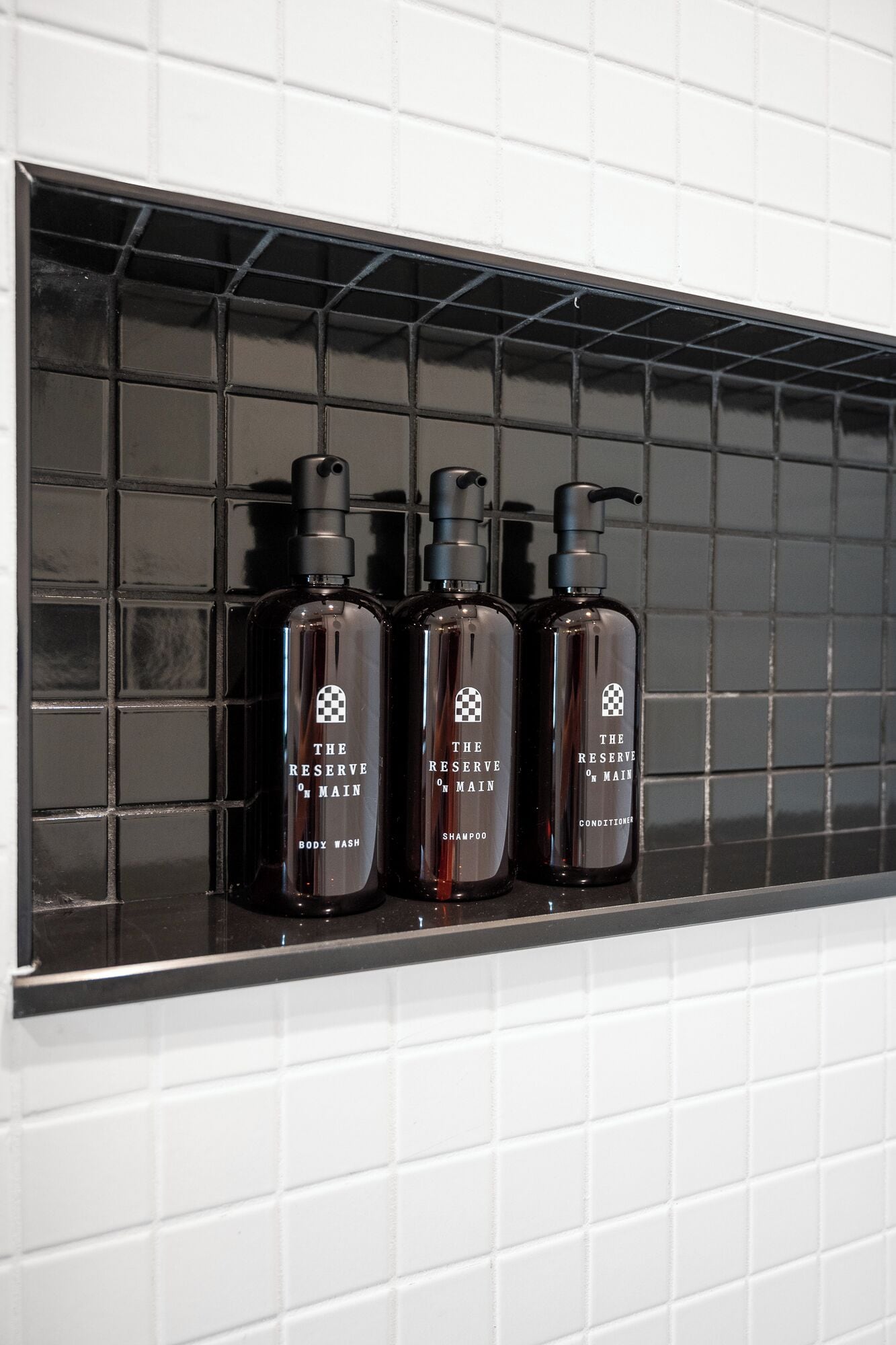 Three bottles of body wash on a black shelf against a white tiled wall.