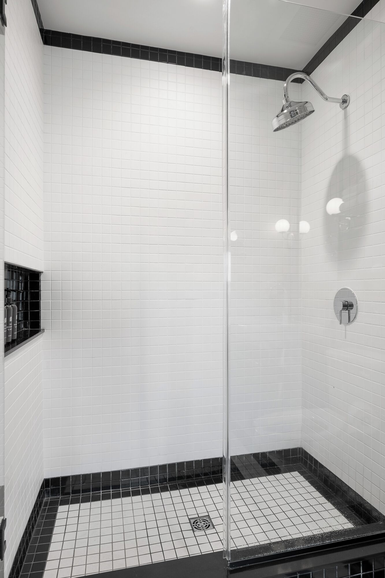 A modern shower with a glass door, white tiled walls, and a black-tiled floor.