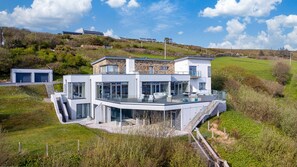 A modern house with a large glass balcony, a stone facade, and a hillside setting.