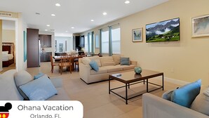 Un salón amplio con sofá, una mesa de centro y un comedor con sillas. Hay ventanas grandes y un cuadro en la pared.