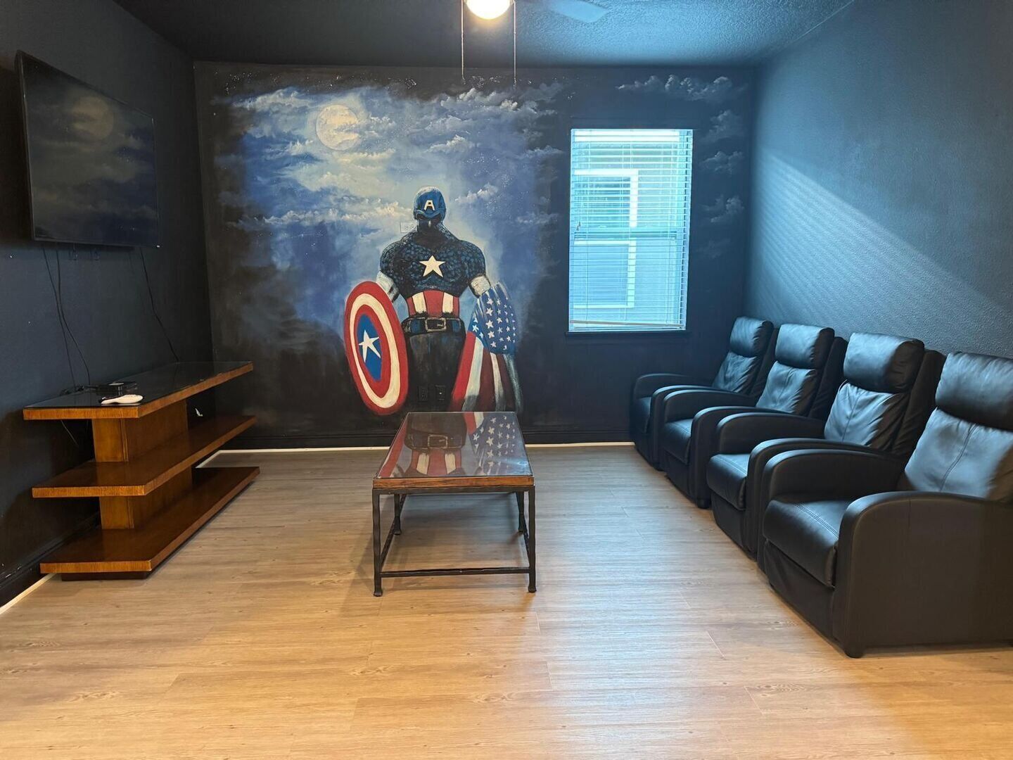 Une pièce avec un plancher en bois, un canapé en cuir noir, une table basse en bois et une fresque murale représentant Captain America.