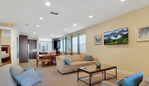 Een ruime woonkamer met een bank, salontafel en een eethoek met stoelen. Er zijn grote ramen en een schilderij aan de muur.