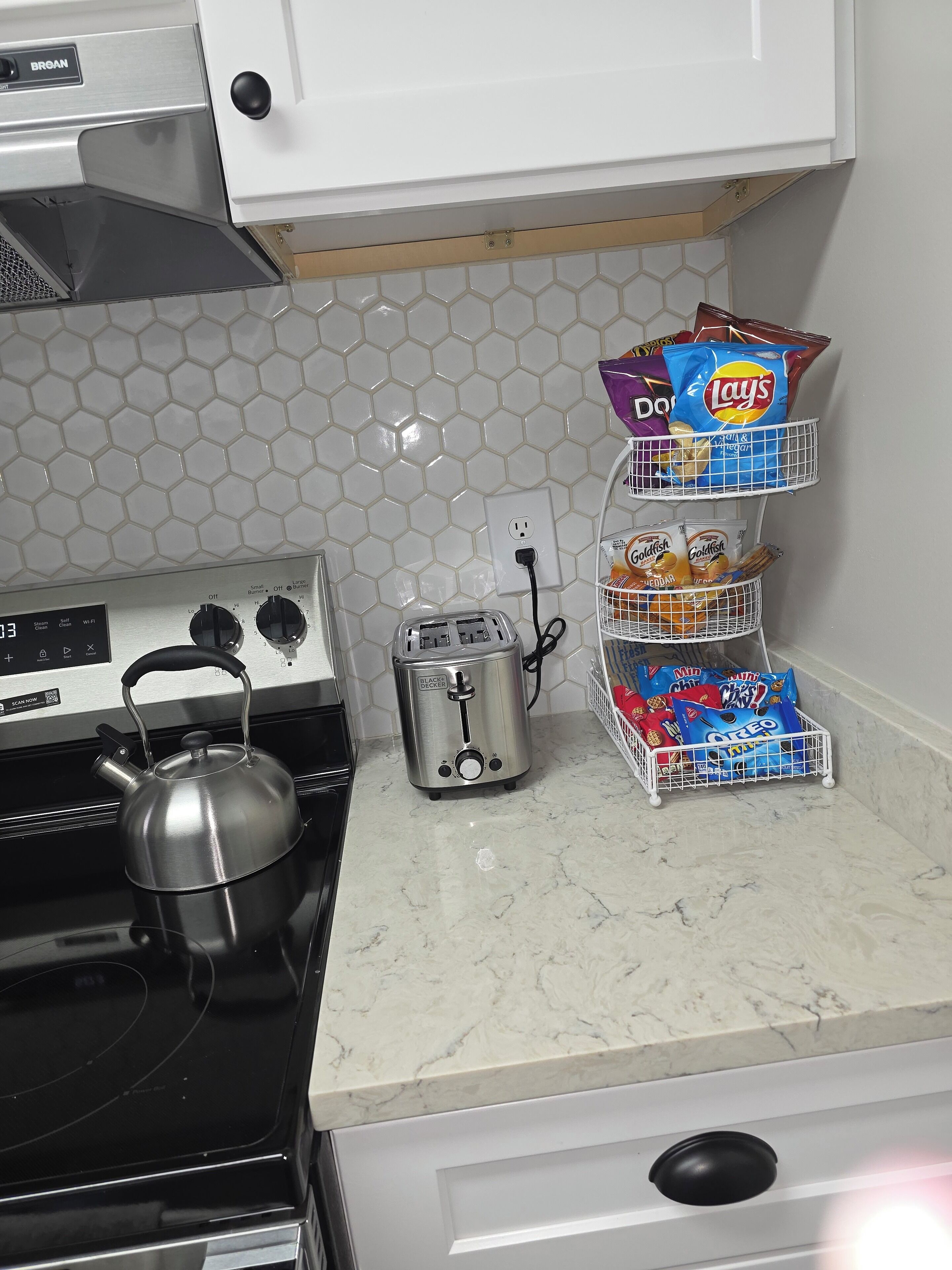 Una cocina con estufa, hervidor, tostadora y un mueble para aperitivos con papas fritas y galletas.