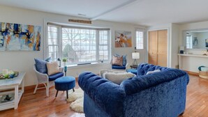Een ruime woonkamer met een groot raam, een blauwe bank, een witte fauteuil en een eettafel met decoratieve objecten.
