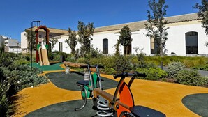 A playground with a slide and swings, surrounded by greenery and a building with a white facade and arched windows. - La Solera de Jerez - Ole Solutions (Jerez de la Frontera)
