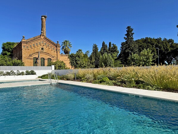A swimming pool with a clear blue water surface, surrounded by a white poolside area. In the background, there is a brick building with a chimney, and a row of tall trees.