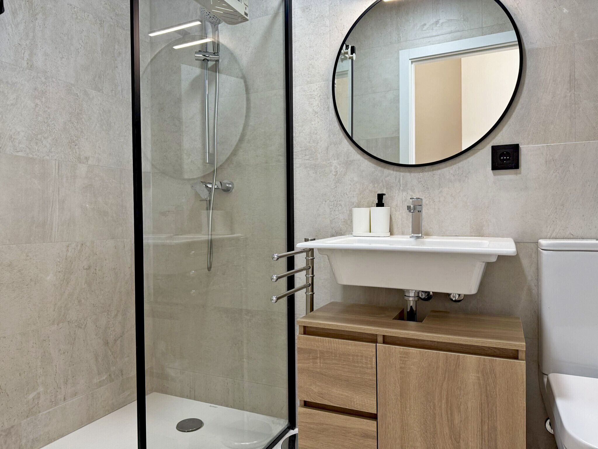 Een moderne badkamer met een douchecabine van glas, een ronde spiegel, een witte wasbak en een houten kastje.