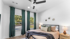 Quarto com cama, mesas de cabeceira, ventoinha no teto e janela com cortinas.