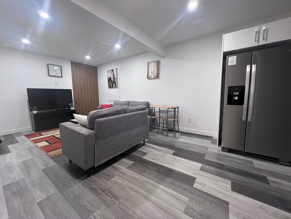 Ein modernes Wohnzimmer mit grauem Sofa, Fernseher, Esstisch und Kühlschrank.