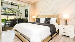 A modern bedroom with a large bed, two bedside lamps, a nightstand, and a sliding glass door leading to an outdoor patio with a table and chairs.