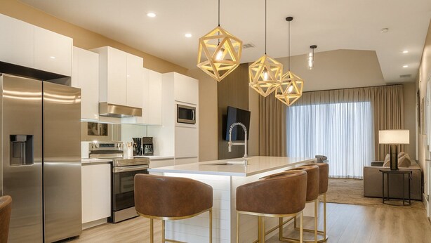 Eine moderne Küche mit Edelstahlgeräten, einer zentralen Kochinsel mit Barhocker, Pendelleuchten und einem Wohnbereich mit Sofa und Lampe.