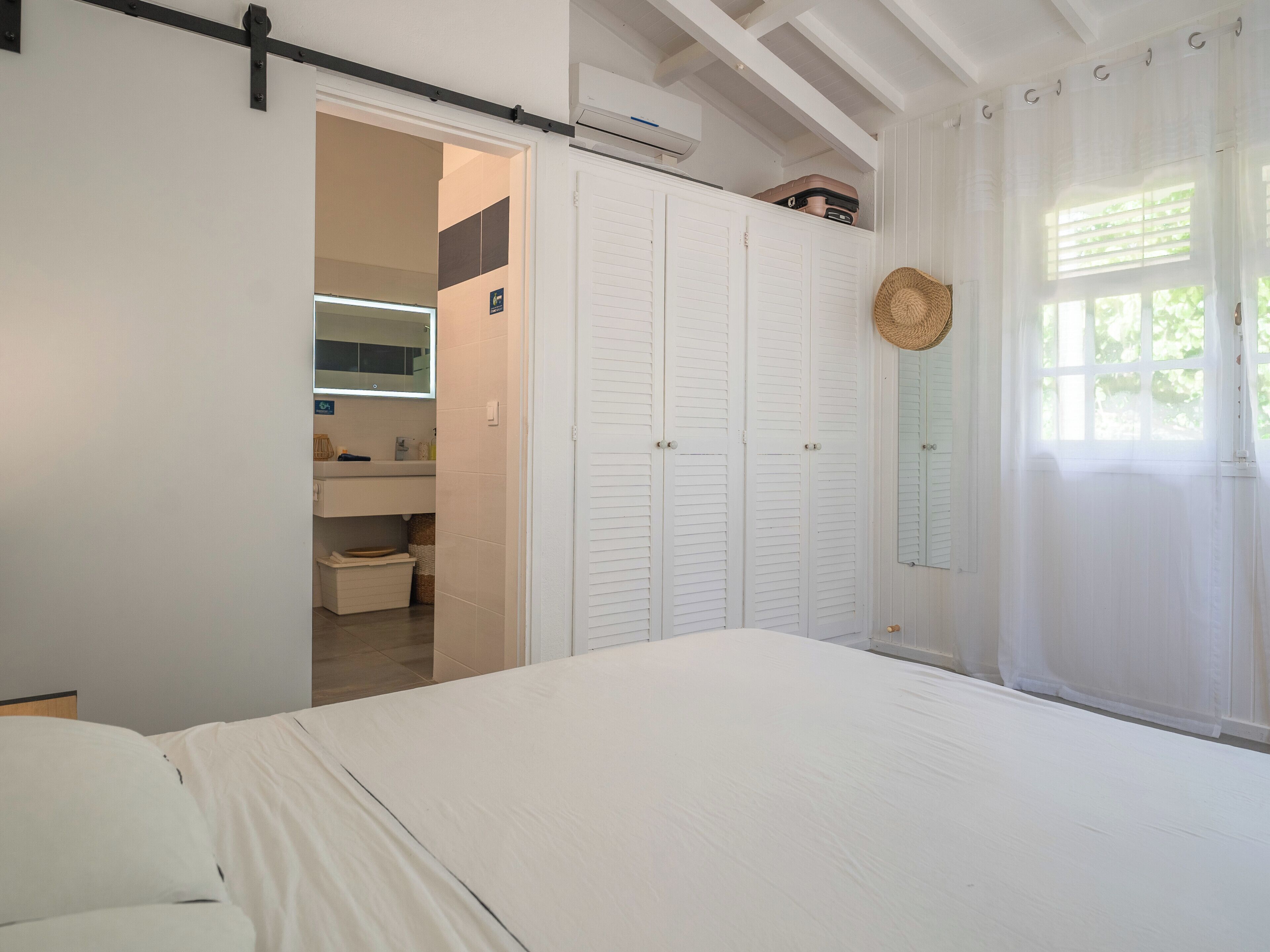 Ein Schlafzimmer mit Bett, Schrank, Badezimmer mit Spiegel und Fenster mit Vorhängen.