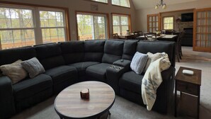 A spacious living room with a large sectional sofa, a round coffee table, and a kitchen area visible through an open door. - Chickadee Retreat - Creekside Pocono Forest, fireplace, games, pool, lakes (Albrightsville)