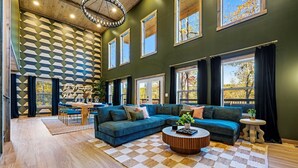 Ein modernes Wohnzimmer mit einem blauen Ecksofa, einem runden Couchtisch und einem karierten Teppich. Mehrere Fenster mit Vorhängen und ein Essbereich mit Tisch und Stühlen sind im Hintergrund zu sehen.