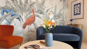 Et værelse med en blå sofa, en orange lænestol, et rundt bord med et magasin, en vase med blomster, et vægmaleri og et indrammet billede.