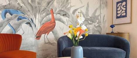 A room with a blue sofa, an orange chair, a round table with a magazine, a vase with flowers, a wall mural, and a framed picture.