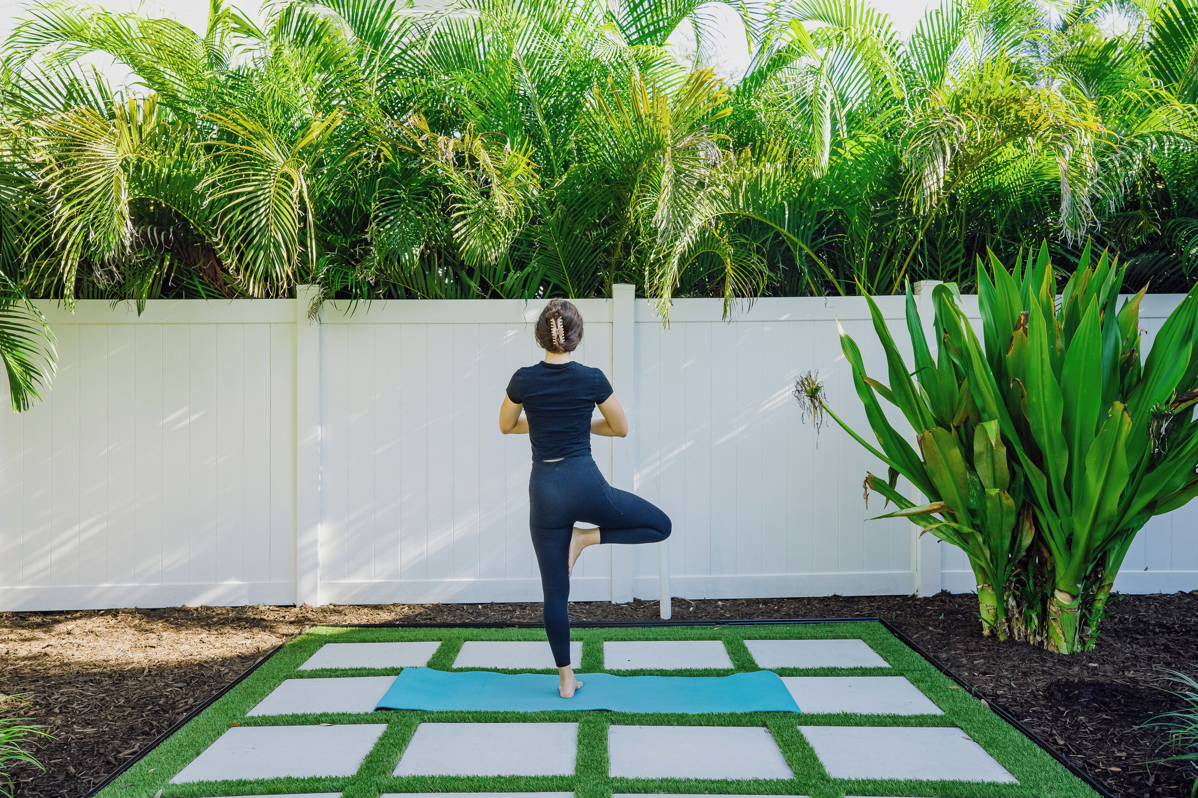 Eine Person übt Yoga auf einer Matte in einem Außenbereich mit weißem Zaun und üppiger Vegetation im Hintergrund.