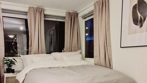 Un dormitorio con una cama grande, una mesita de noche con lámpara, una ventana con cortinas y un cuadro en la pared.