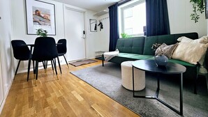 Una sala de estar moderna con un sofá verde, una mesa de centro redonda y sillas negras. Hay una ventana con cortinas oscuras, un cuadro en la pared y una planta colgando del techo.