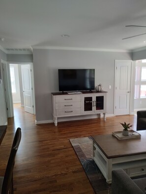 A living room with a flat-screen TV on a white cabinet, wooden flooring, and a dining area with a table and chairs.
