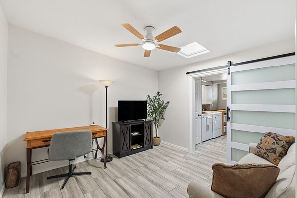 A modern living room with a wooden desk, a grey office chair, a TV on a black cabinet, a ceiling fan, a potted plant, and a sliding door leading to a kitchen. - Prime Central Location- Minutes from TMC Hospital: Desert House (Tucson)