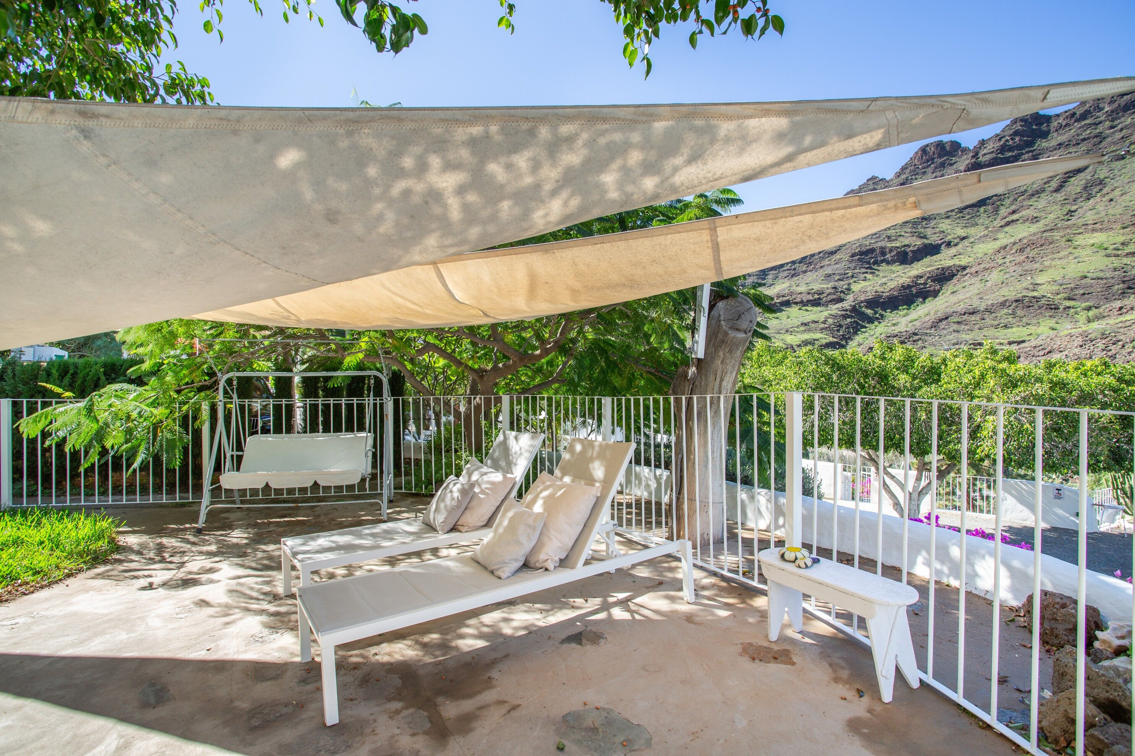 A shaded outdoor seating area with white lounge chairs and a bench, surrounded by a white fence, with a mountainous backdrop.