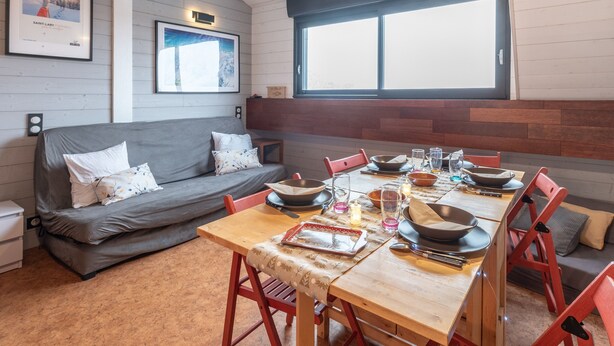 A cozy room with a wooden dining table set for four, a grey sofa, and a large window.