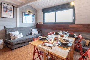 Um quarto aconchegante com uma mesa de jantar de madeira posta para quatro pessoas, um sofá cinza e uma janela grande.
