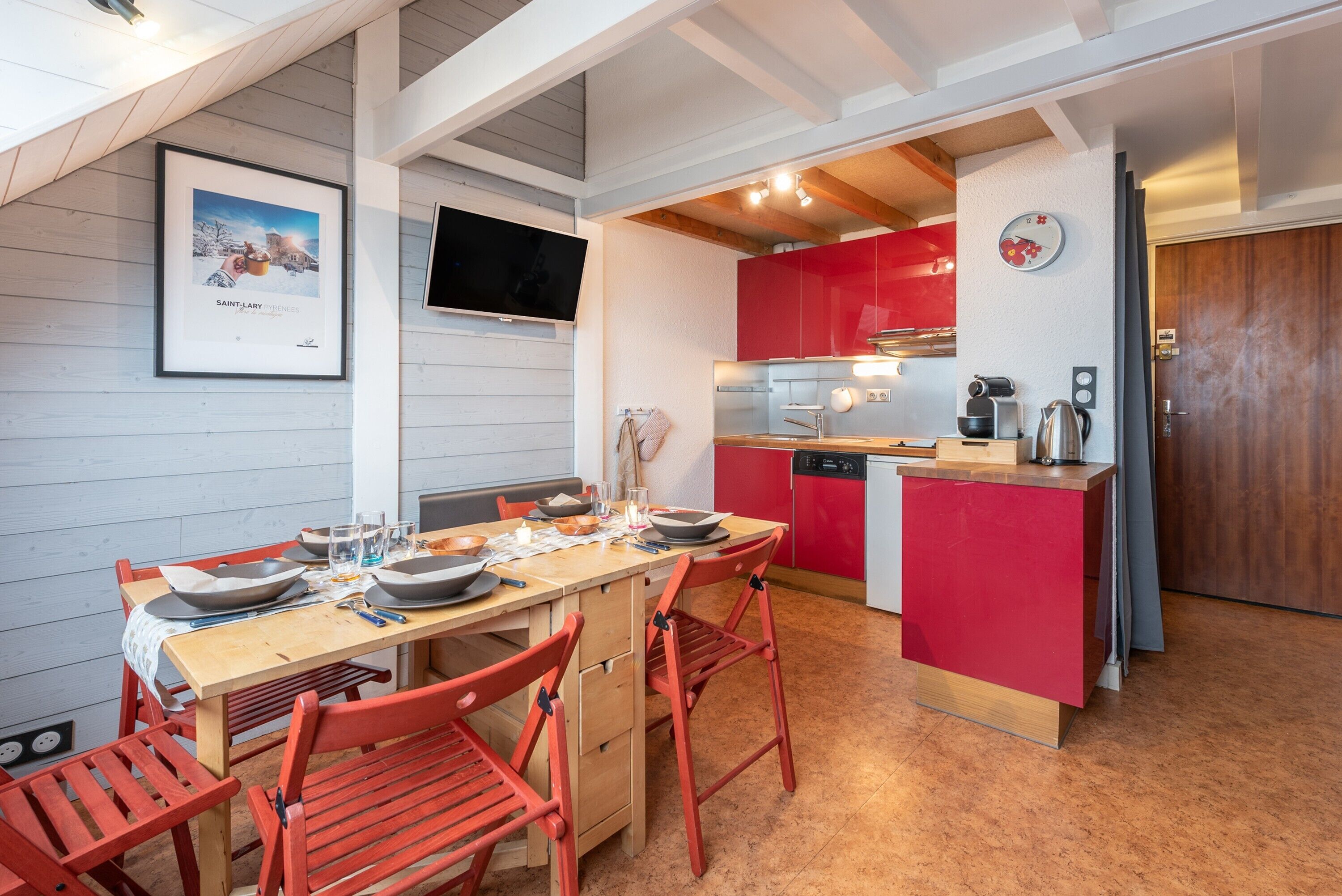 Una cocina moderna con armarios rojos, una mesa de comedor de madera con sillas y un televisor montado en la pared.