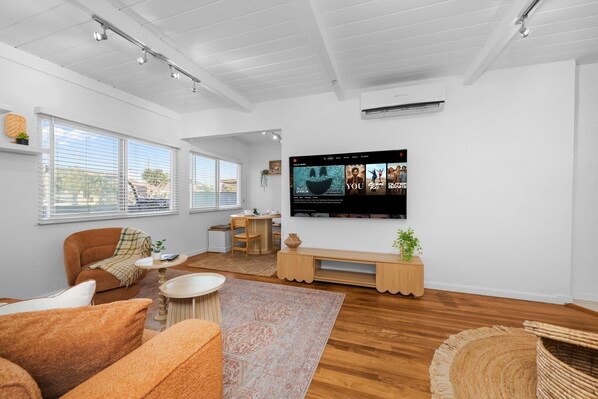 Un salon moderne avec un téléviseur à écran plat fixé au mur, une table basse en bois, un canapé et un coin repas comprenant une table et des chaises en bois.