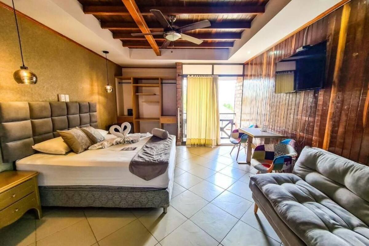 A modern hotel room with a large bed, a sofa, a dining area, and a ceiling fan.