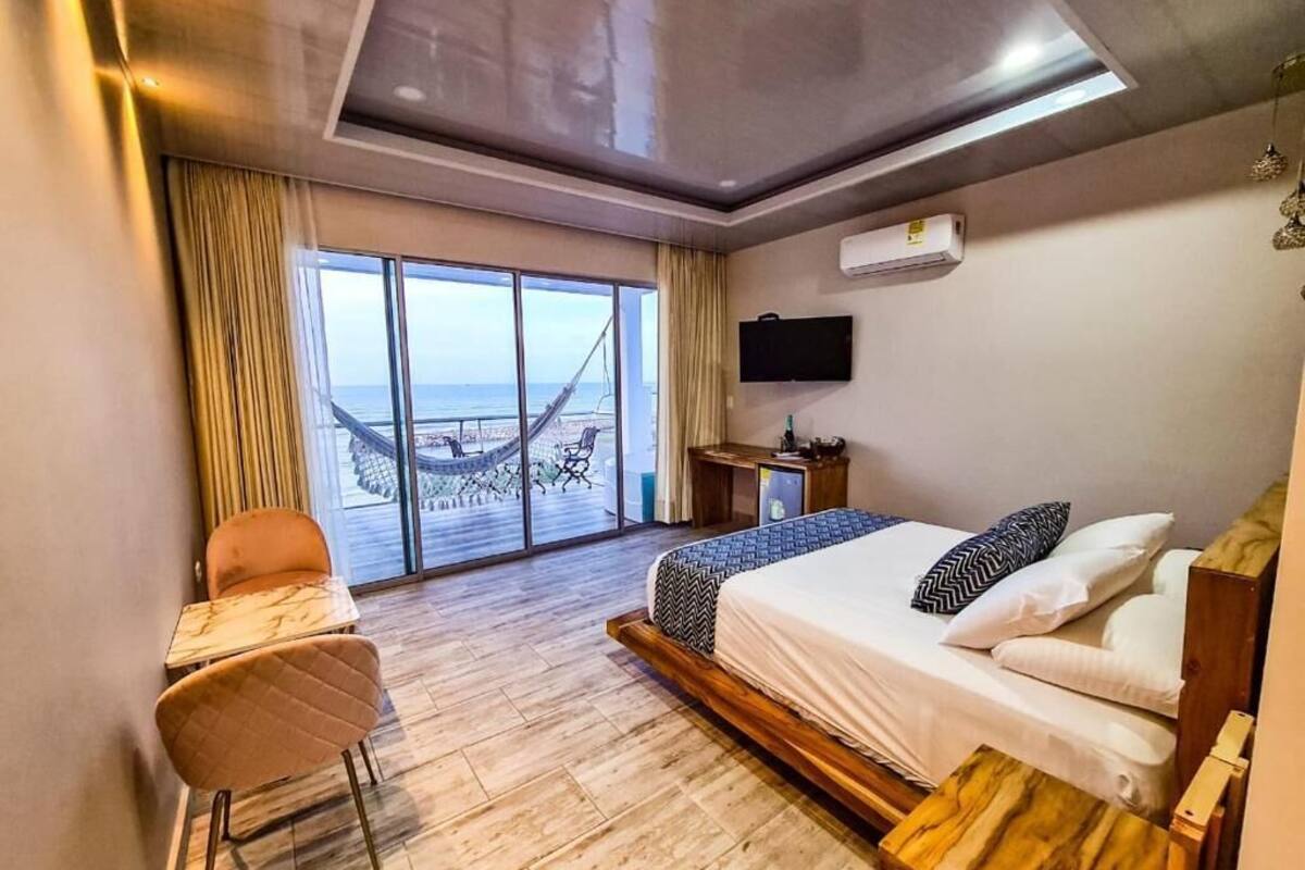 A hotel room with a bed, a desk, a chair, and a large window with a view of the beach.