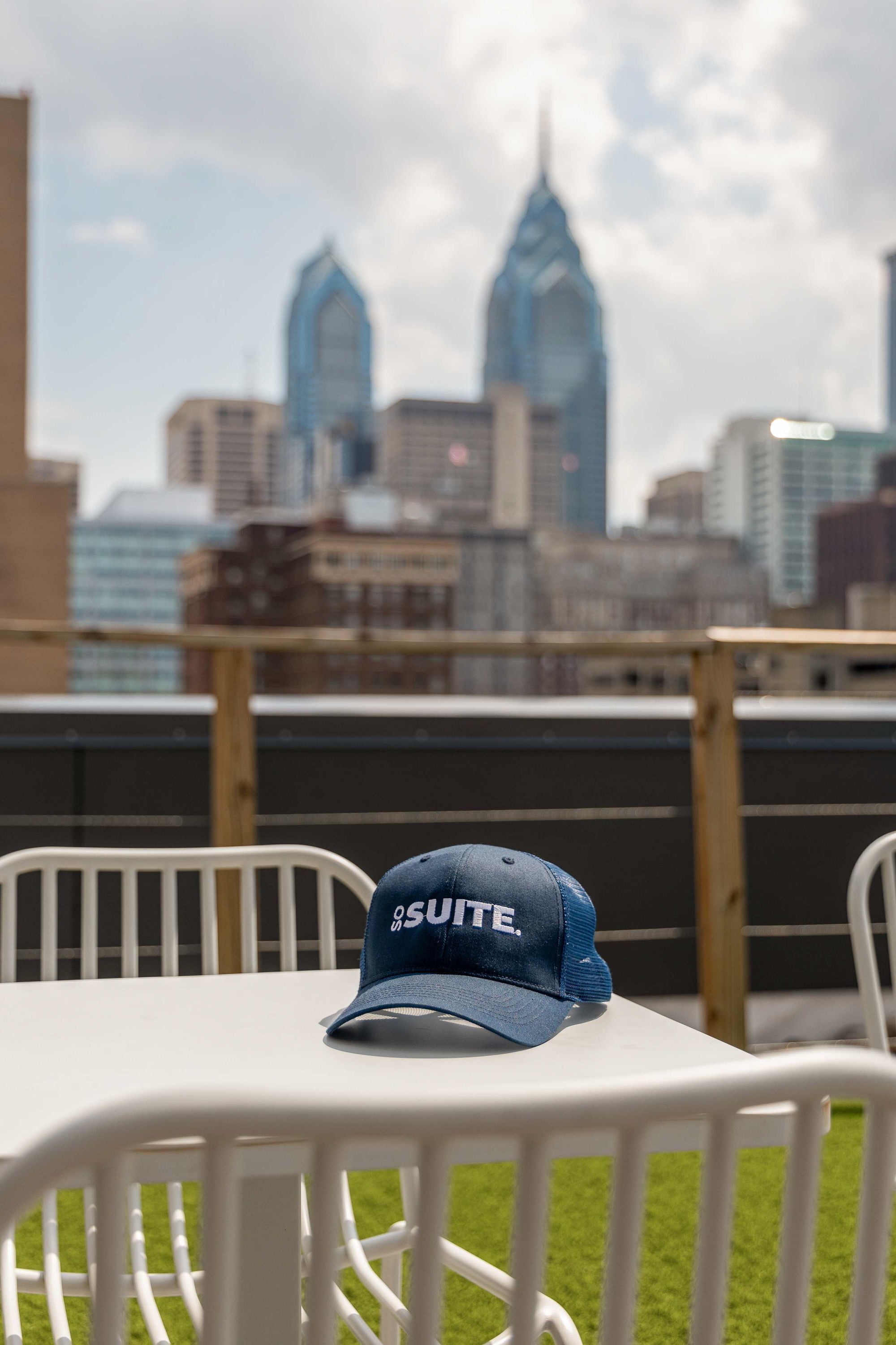 A rooftop terrace with a view of a city skyline, including a distinctive building with a spire. A blue cap with the word "SUITE" is placed on a white table.
