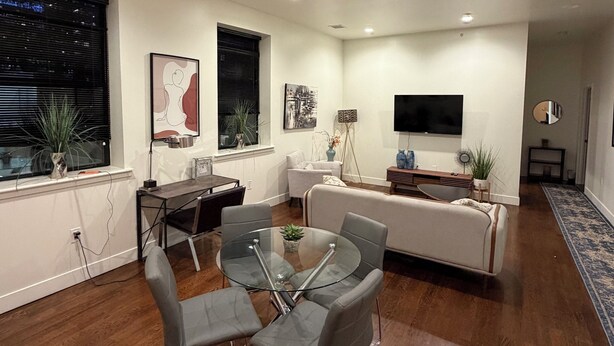 Ein modernes Wohnzimmer mit einem Esstisch mit Glasplatte, grauen Polsterstühlen, einem weißen Sofa und einem flachbildfernseher an der Wand.