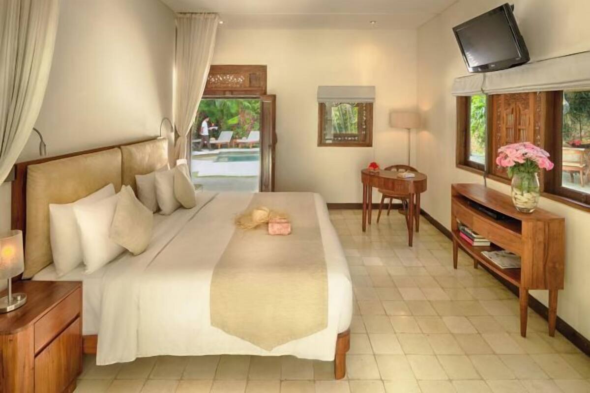 A hotel room with a bed, a TV, a small table, a chair, a window with a view of a pool, and a wooden cabinet.