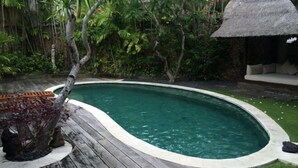 A swimming pool with a wooden deck, surrounded by lush greenery and a thatched-roof structure. - Villa Kubu Seminyak (Bali)
