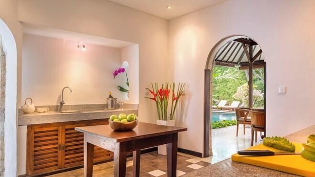 A kitchen with a granite countertop, wooden cabinets, a dining table, and a view of an outdoor pool area. - Villa Kubu Seminyak (Bali)