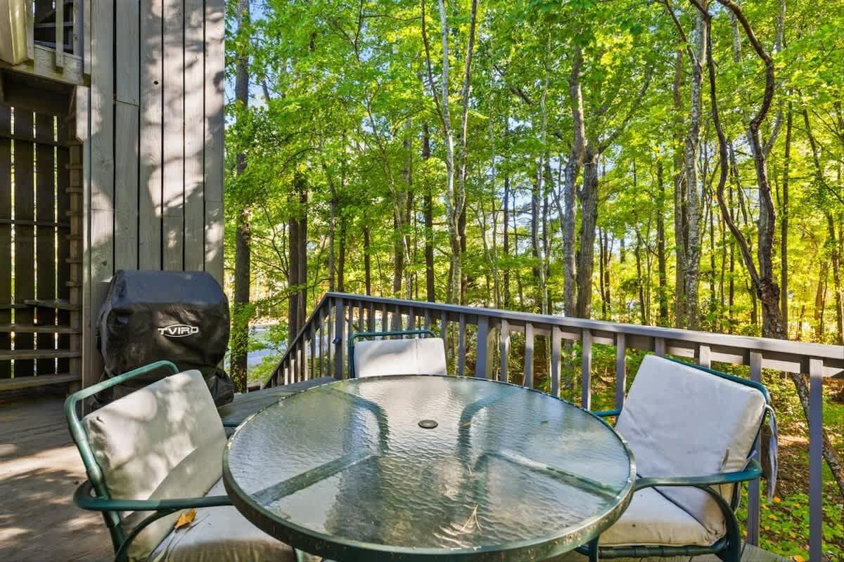 Un patio al aire libre con una mesa redonda de vidrio y dos sillas, rodeado por una terraza de madera y una barandilla de metal.