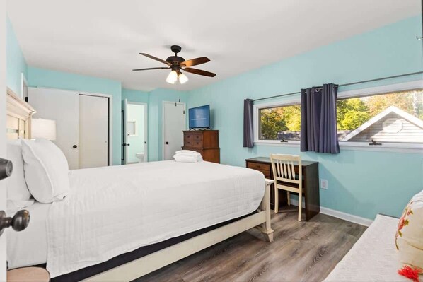 Une chambre à coucher comprenant un lit, un bureau, une chaise, un ventilateur de plafond et une fenêtre avec des rideaux.