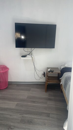 Une chambre avec un téléviseur à écran plat fixé au mur, une table de chevet en bois avec une lampe, un lit recouvert d’une courtepointe à motifs blancs et bleus, et un panier à linge rose posé sur le sol.