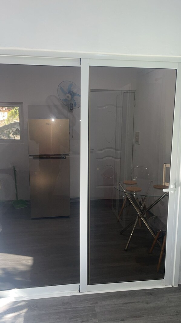 Une cuisine équipée d’un réfrigérateur en acier inoxydable, une porte vitrée donnant sur un coin repas avec une table et des chaises en verre, et un ventilateur fixé au mur.
