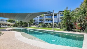 A swimming pool with a curved edge, surrounded by a landscaped area with palm trees and a building with balconies in the background. - Pavillions  Ocean Breeze Apartment 119 (Agnes Water)