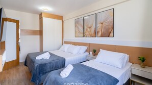Une chambre d’hôtel avec deux lits, une armoire en bois, une table de chevet avec une plante et un mur orné de trois œuvres d’art encadrées.